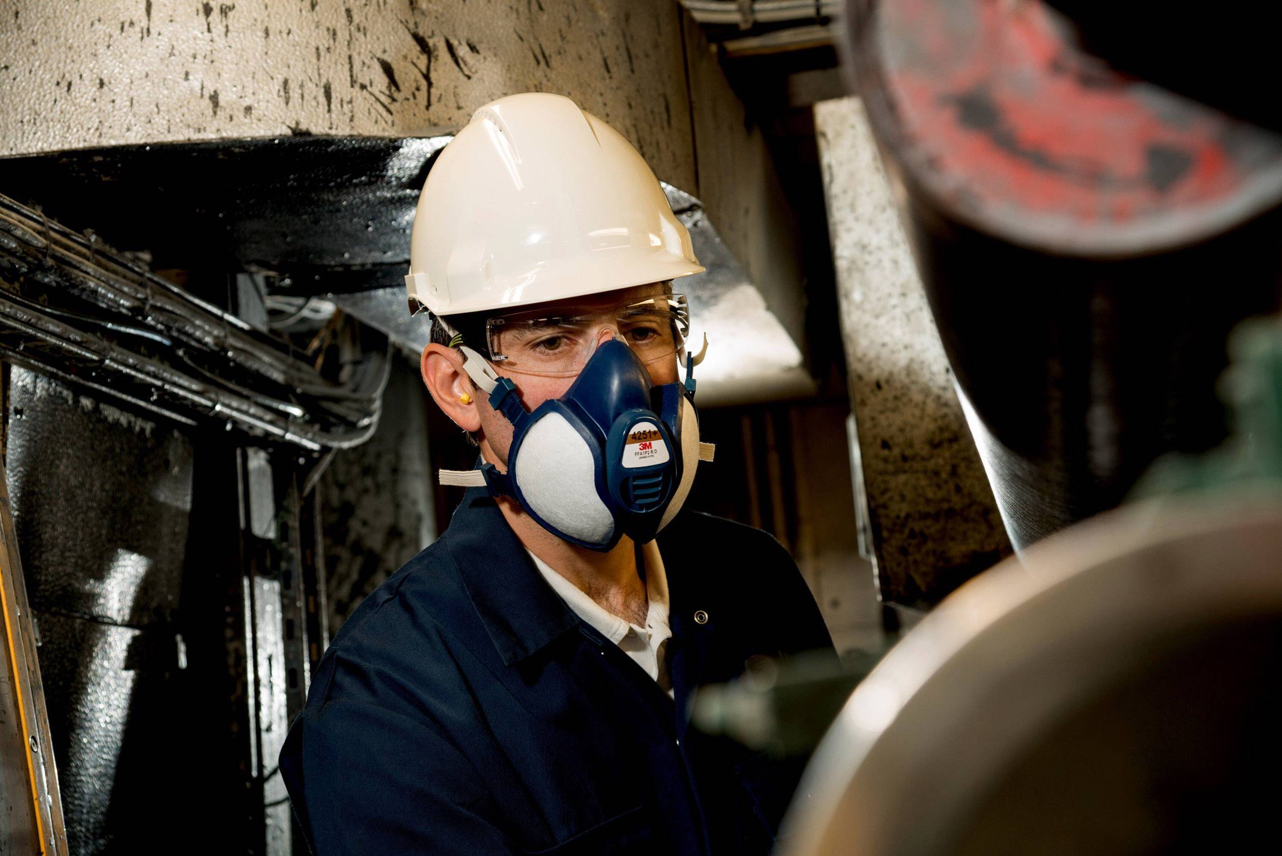 Eine Person trägt Schutzkleidung und eine Atemmaske in einer industriellen Umgebung, möglicherweise bei der Arbeit mit Maschinen.