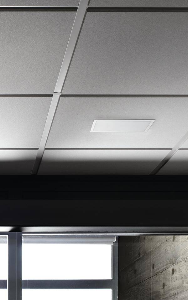 Abgehängte Decke mit quadratischen Deckenkacheln und integriertem Lüftungsgitter über einem Fenster. Holzverkleidete Wand sichtbar.