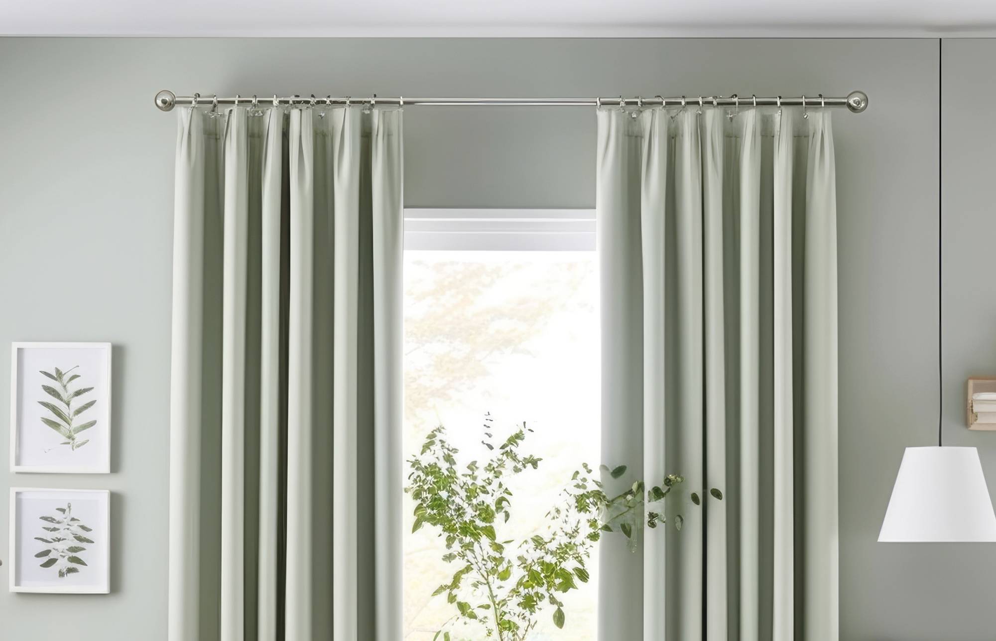 Hellsalbeigrüne Vorhänge rahmen ein helles Fenster, mit gerahmten botanischen Drucken auf der linken Seite und einer Stehlampe auf der rechten Seite in einem ruhigen Raum.
