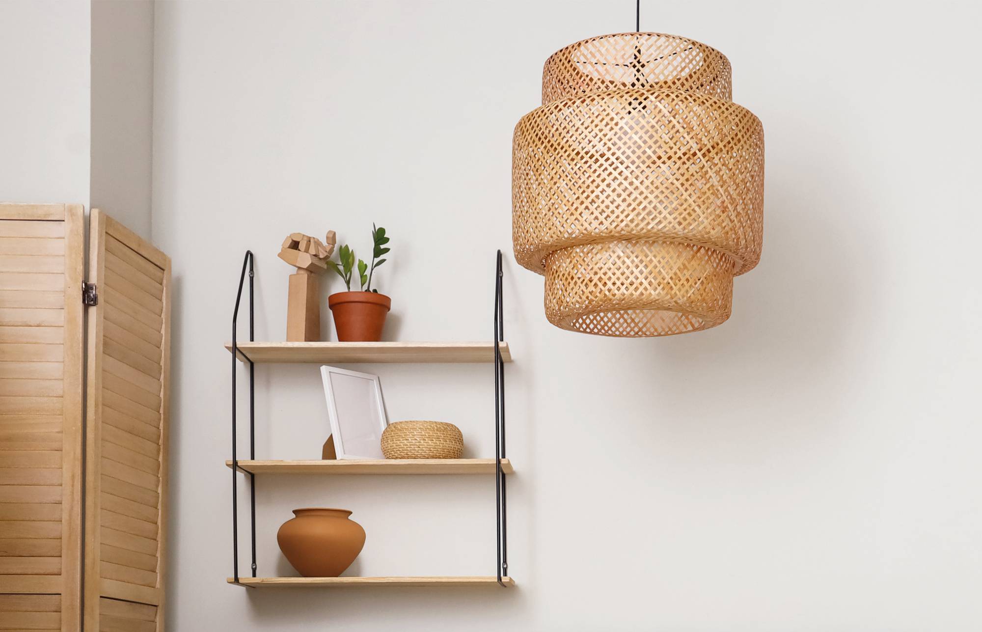 Minimalistisches Raumeck mit einer geflochtenen Pendelleuchte über einem Wandregal, das eine Pflanze, einen Rahmen, einen Korb und Keramik hält, neben einem Holzraumteiler.