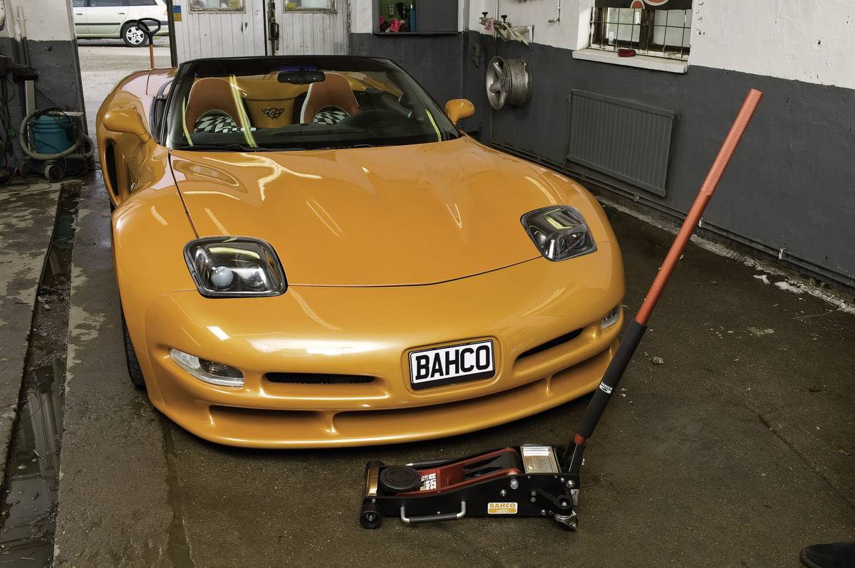 Orangefarbener Sportwagen in Werkstatt mit Wagenheber davor. Nummernschild zeigt 'BAHCO'.