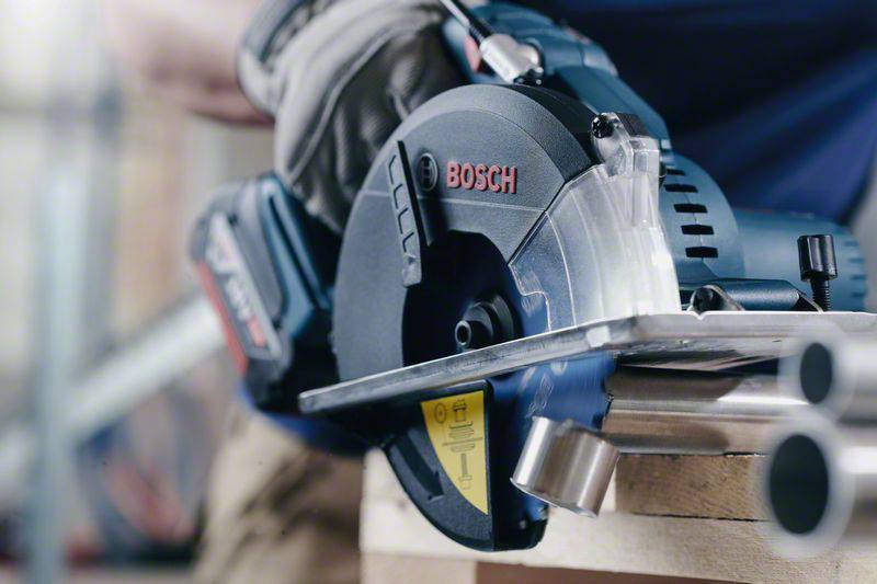 Eine Person schneidet Metallrohre mit einer elektrischen Kreissäge. Die Säge hat das Bosch-Logo. Sie trägt Handschuhe für Sicherheit.