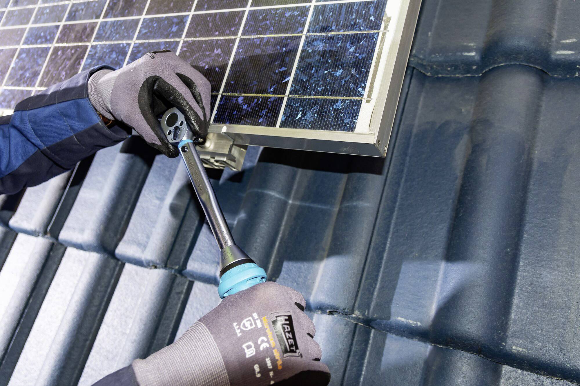 Eine Person installiert ein Solarpanel auf einem Dach. Sie hält ein Werkzeug in Handschuhen und befestigt das Panel an der Halterung.