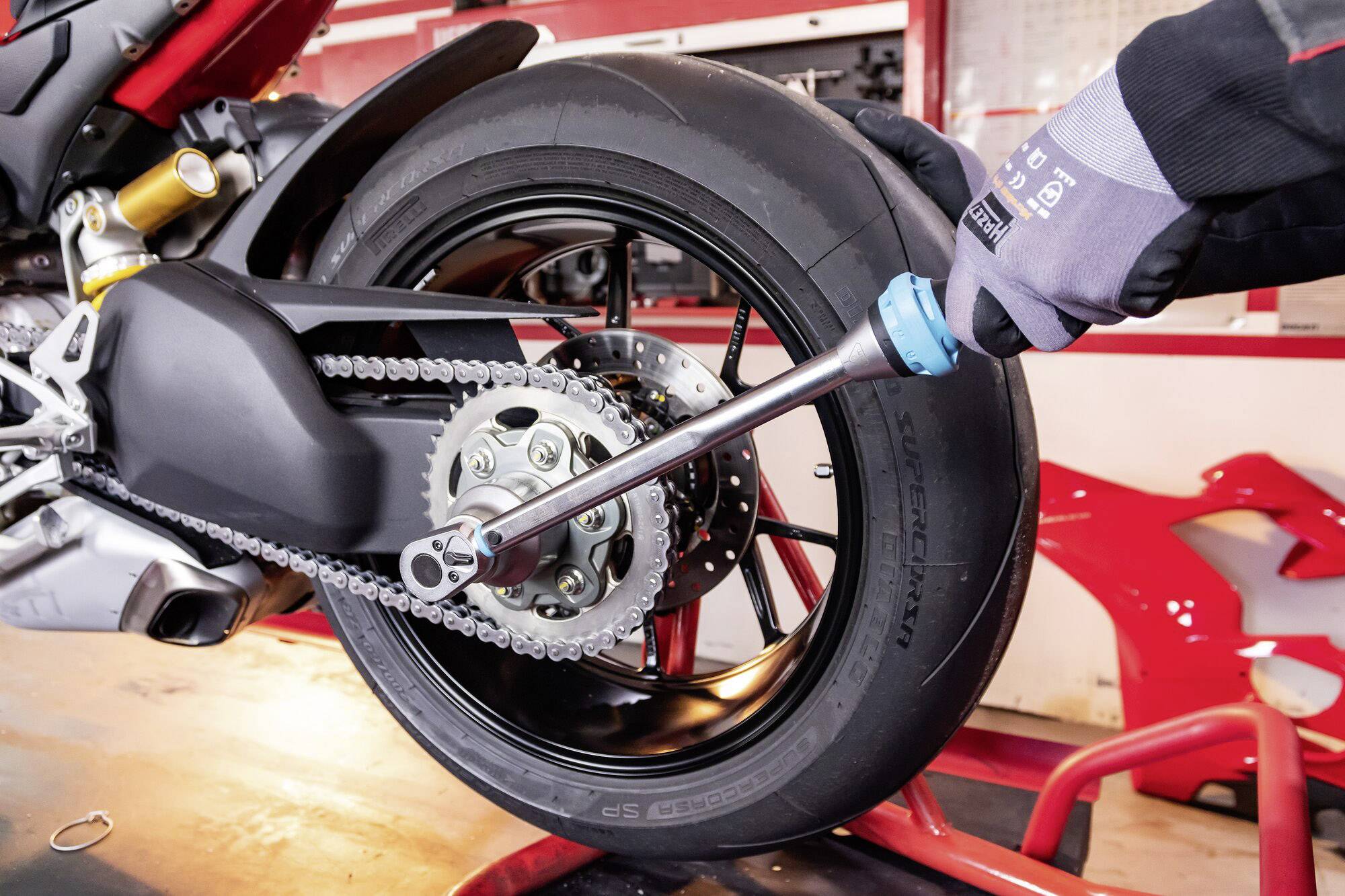 Werkzeug wird verwendet, um an einem Motorrad das Hinterrad zu justieren. Techniker trägt Handschuhe und arbeitet in einer Werkstattumgebung.