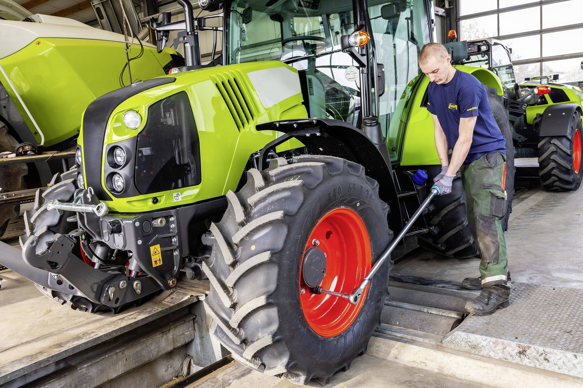 Ein Mechaniker repariert ein großes landwirtschaftliches Fahrzeug in einer Werkstatt. Der Traktor ist grün mit rotem Rad.