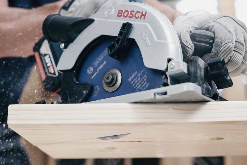 Eine Kreissäge schneidet Holz, während eine Person sie mit Sicherheitshandschuhen führt. Der Fokus liegt auf der Säge und dem Holzbearbeitungsprozess.