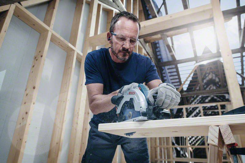 Ein Mann mit Schutzbrille sägt ein Holzstück in einer im Bau befindlichen Holzstruktur. Er trägt Arbeitshandschuhe und wirkt konzentriert.
