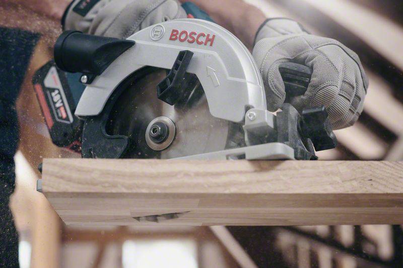 Eine Person schneidet mit einer Handkreissäge ein Holzbrett. Die Säge ist silberfarben und wird von beiden Händen in Arbeitshandschuhen gehalten.