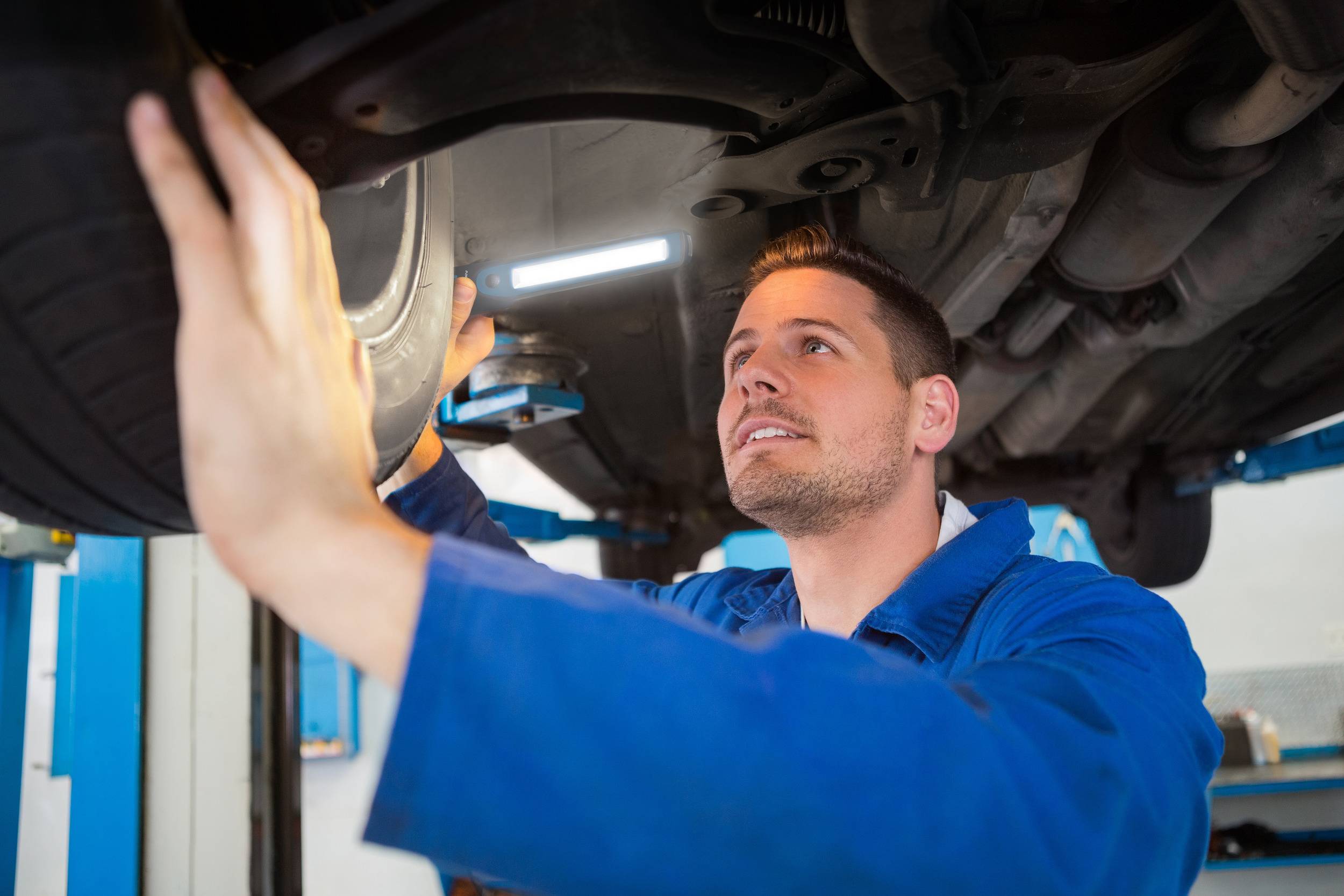 Ein Mechaniker in blauer Arbeitskleidung inspiziert den Unterboden eines Autos auf einer Hebebühne in einer Werkstatt.