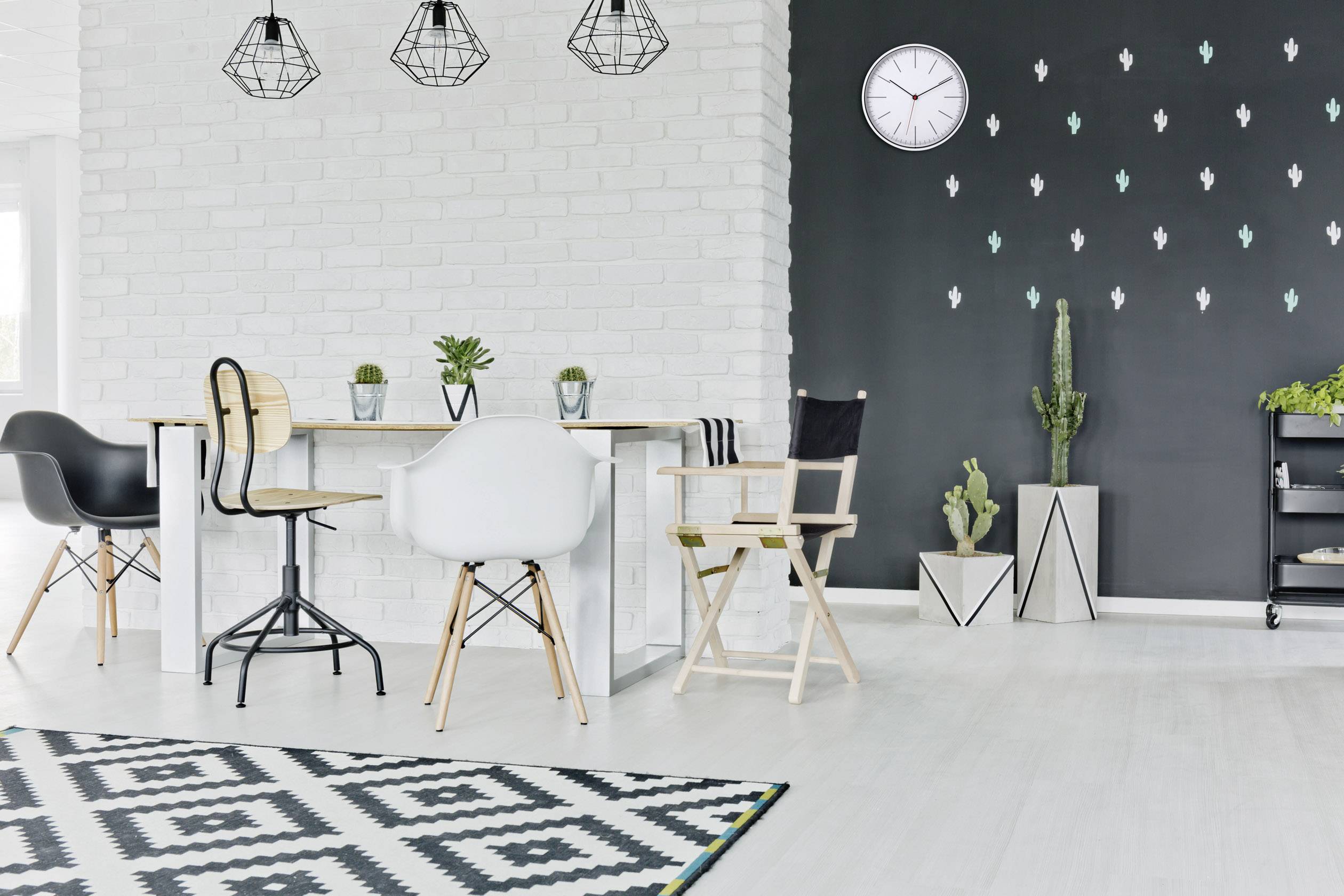 Esszimmer mit hellem Holztisch und verschiedenen Stühlen, weiße Backsteinwand, schwarze Wand mit Uhr und Kaktusdekorationen.