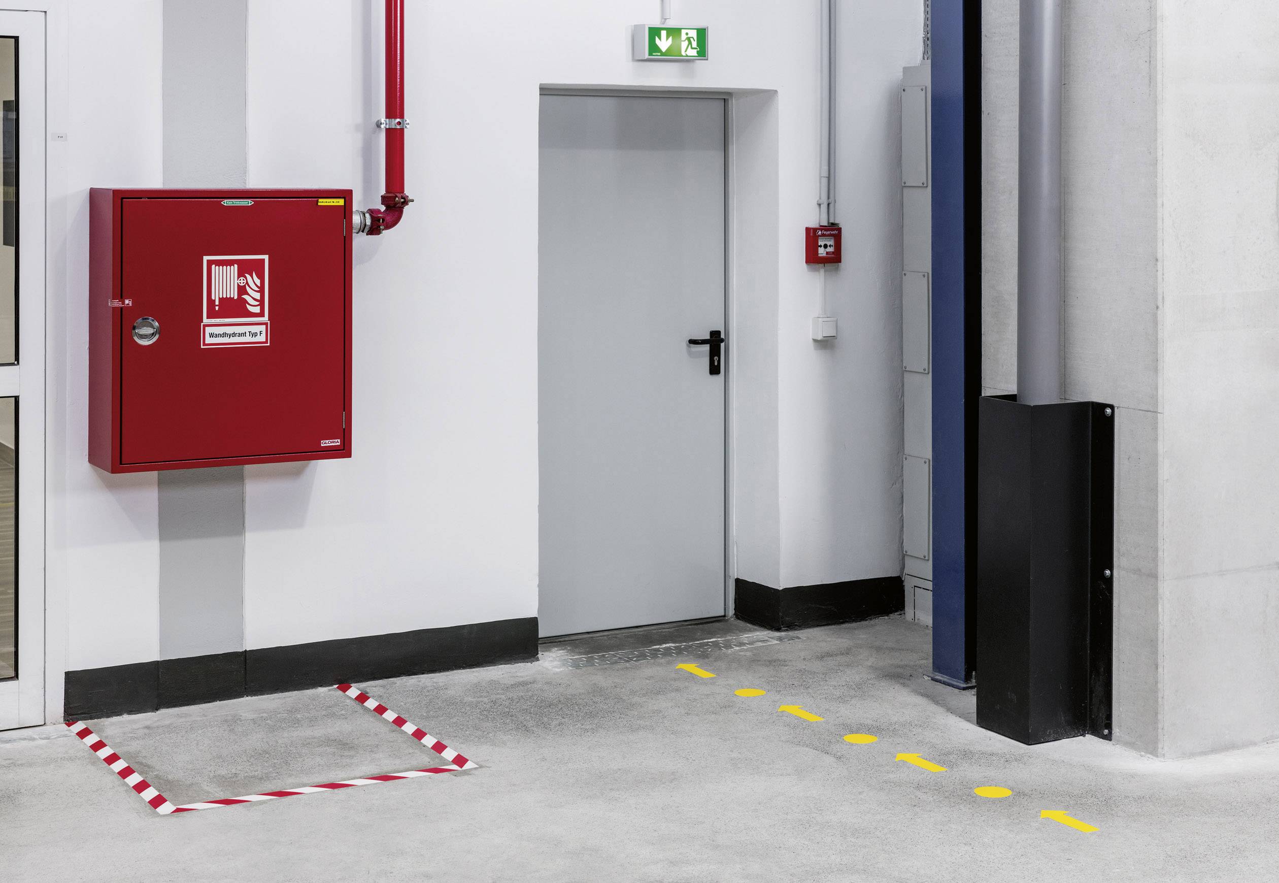 Eine Notausgangstür in einem Lager mit grüner Fluchtschildmarkierung darüber. Daneben ein roter Feuerlöscherkasten an der Wand.