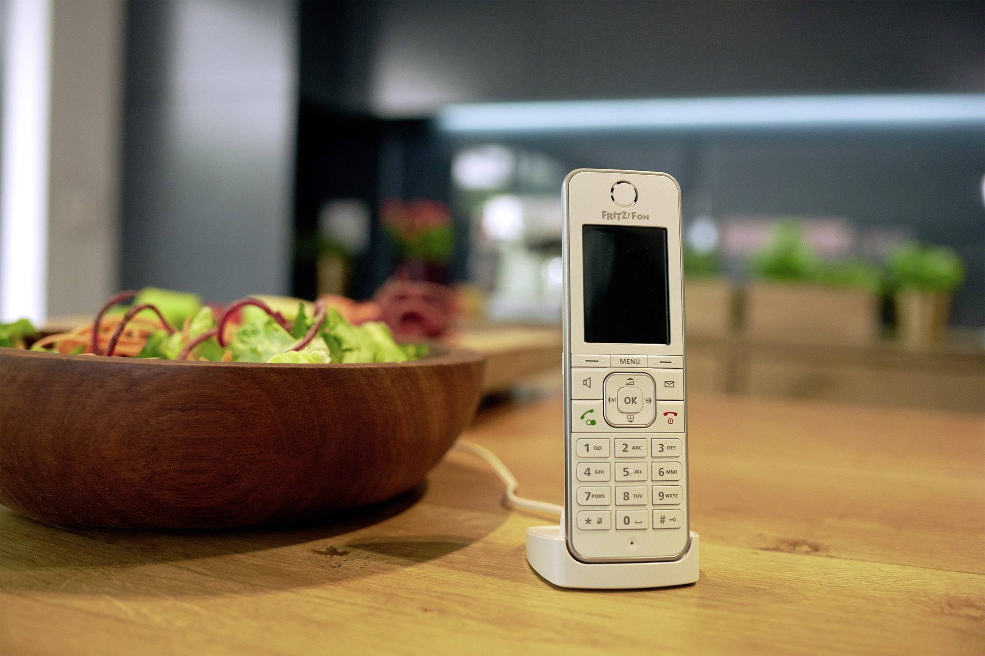 Ein schnurloses Telefon steht auf einem Tisch neben einer Schüssel mit Salat. Im Hintergrund sind verschwommene Küchenelemente sichtbar.