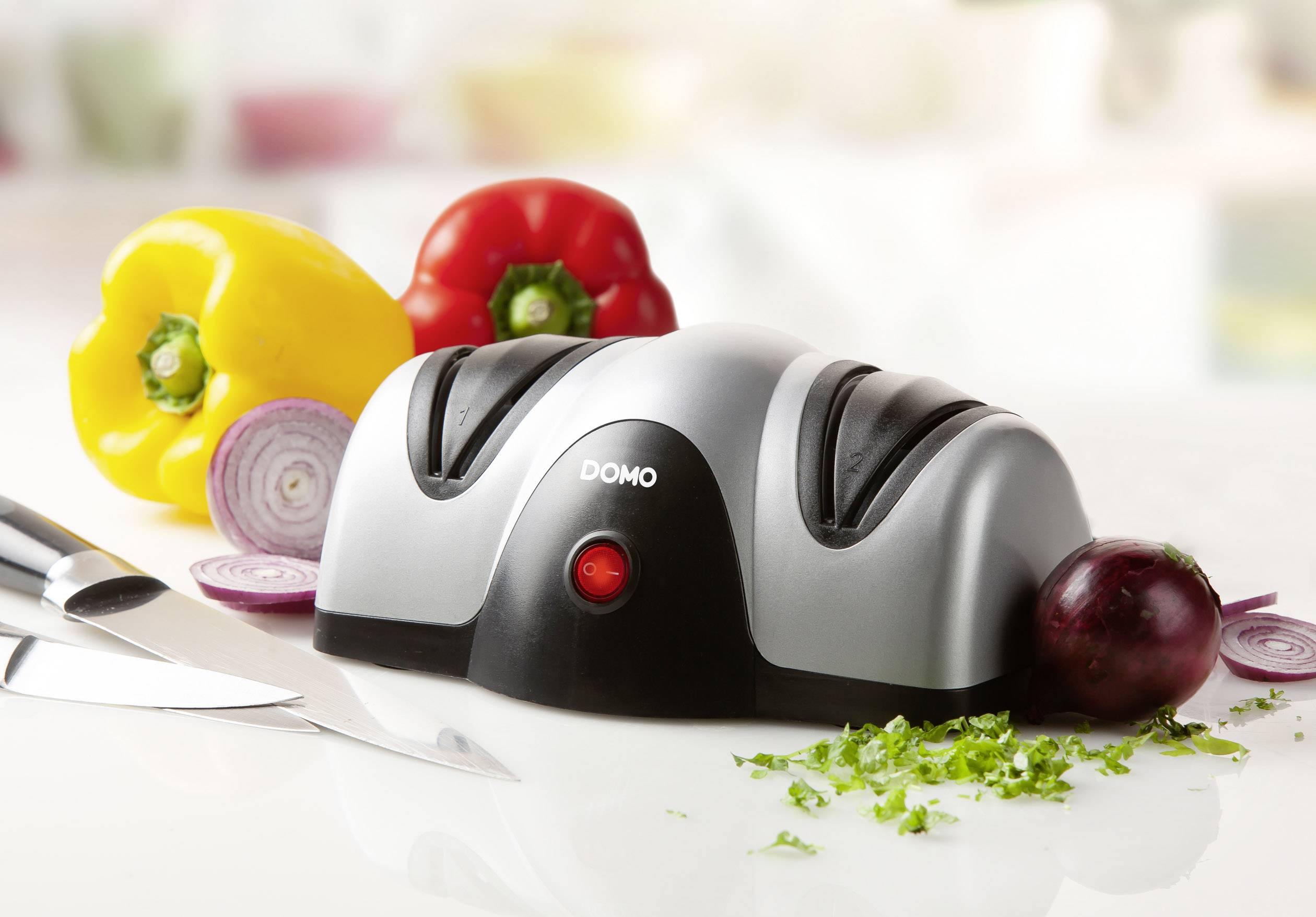 Elektrischer Messerschärfer mit roter Kontrollleuchte auf Küchentisch, umgeben von Paprika, Zwiebeln und einem Messer.