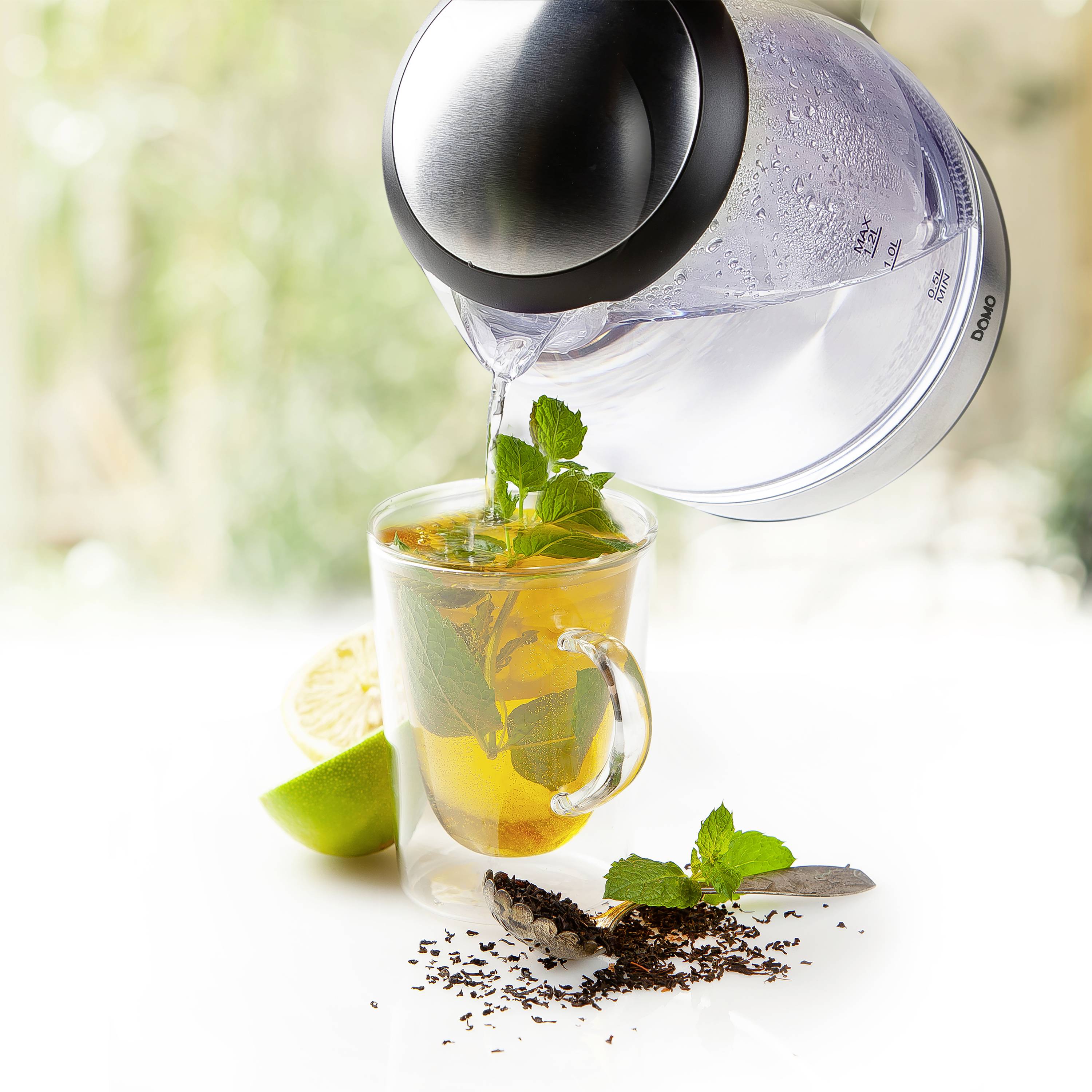 Ein Glas Tee wird mit einem Wasserkocher aufgefüllt. Im Glas befinden sich frische Minzblätter und Zitronenscheiben. Teeblätter und eine Limettenscheibe liegen daneben.
