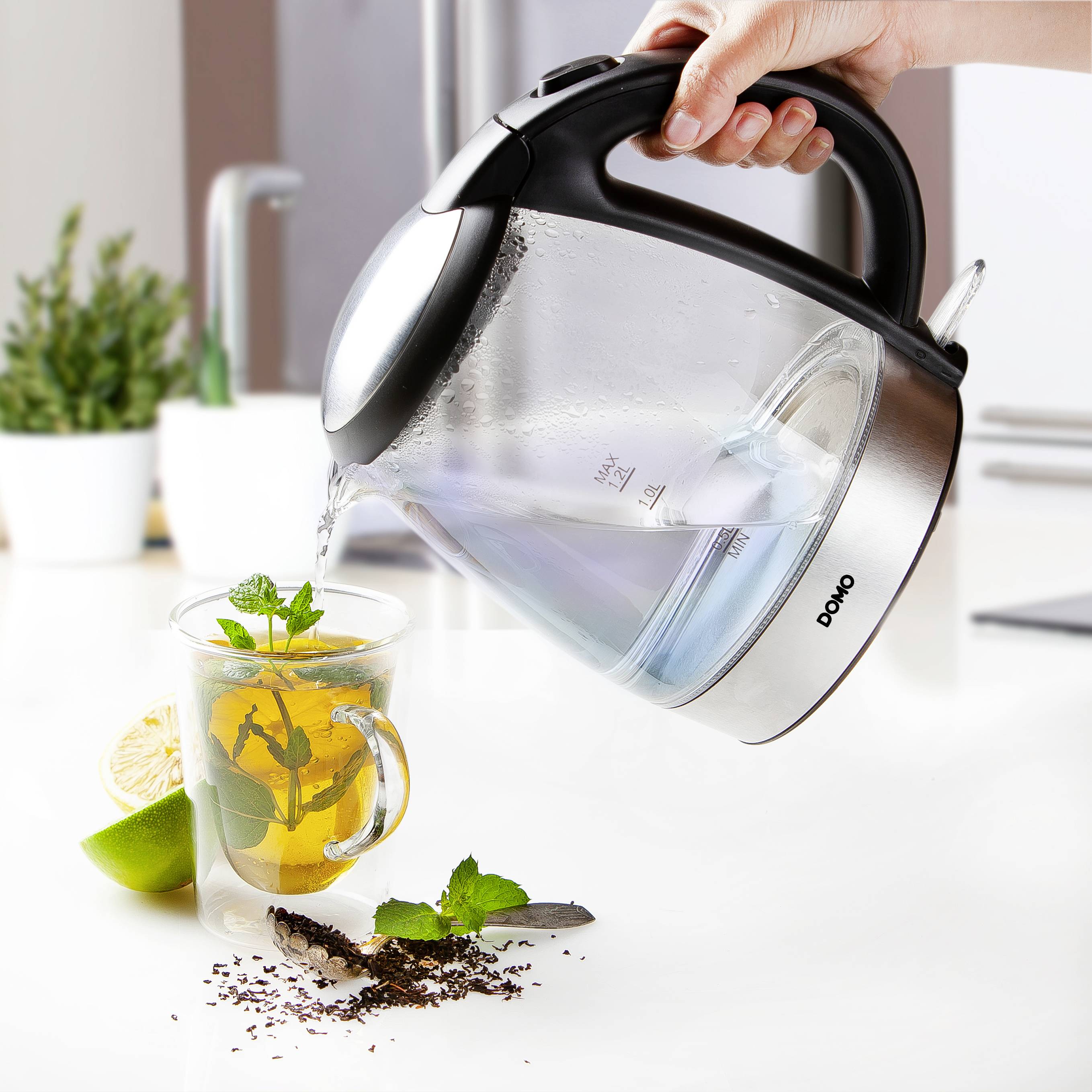 Eine Hand gießt heißes Wasser aus einem Wasserkocher in eine Tasse mit Tee, Minzblättern und Zitronenscheiben neben einer Limette.