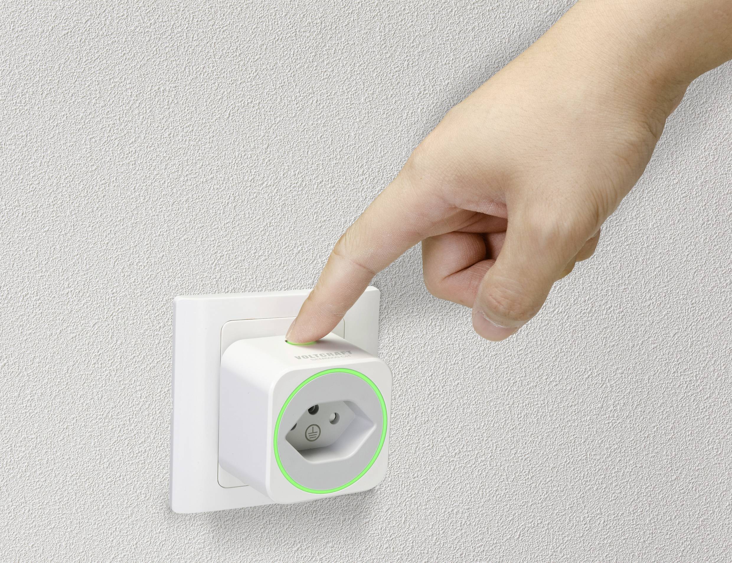 Eine Hand steckt einen smarten Stecker in eine weiße Wandsteckdose, der Stecker leuchtet grün. Der Hintergrund ist beige.