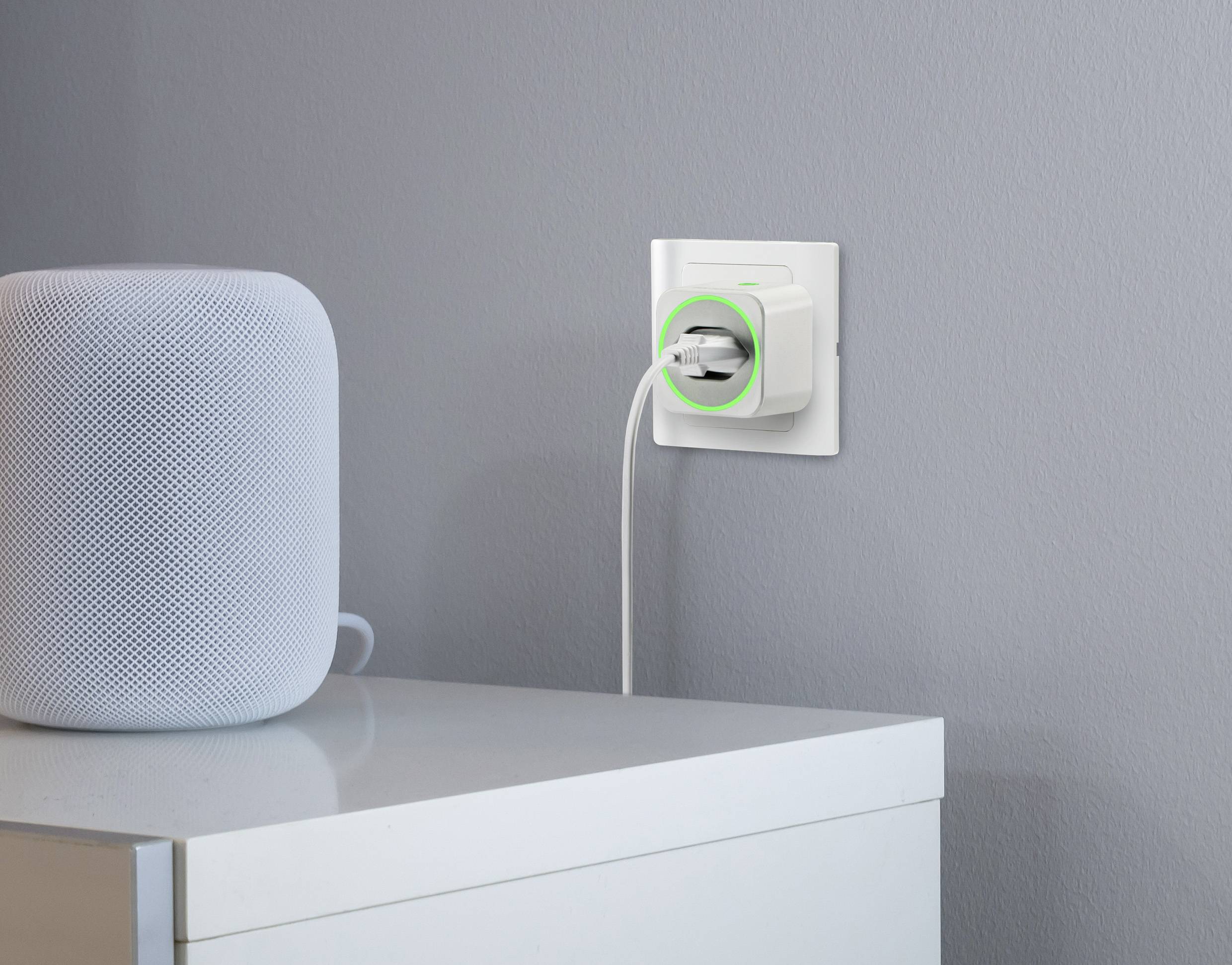 Weiße Steckdose an grauer Wand, ein Smart-Home-Gerät mit grünem Leuchtring. Daneben auf einem weißen Tisch steht ein Lautsprecher.