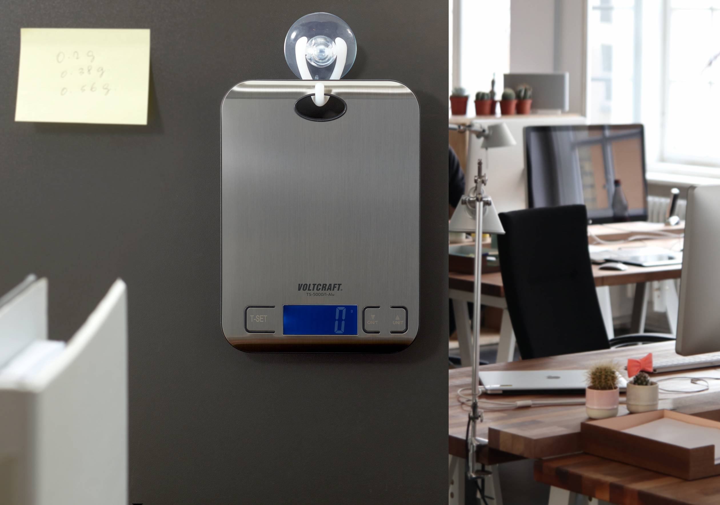 Eine digitale Küchenwaage hängt mit einem Saughaken an einer Wand in einem Büro mit modernen Schreibtischen und Computern.