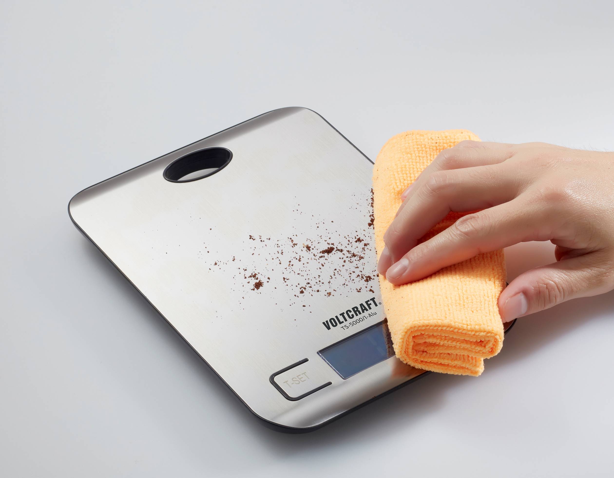 Hand wischt Kaffeepulver von digitaler Küchenwaage mit gelbem Tuch ab.