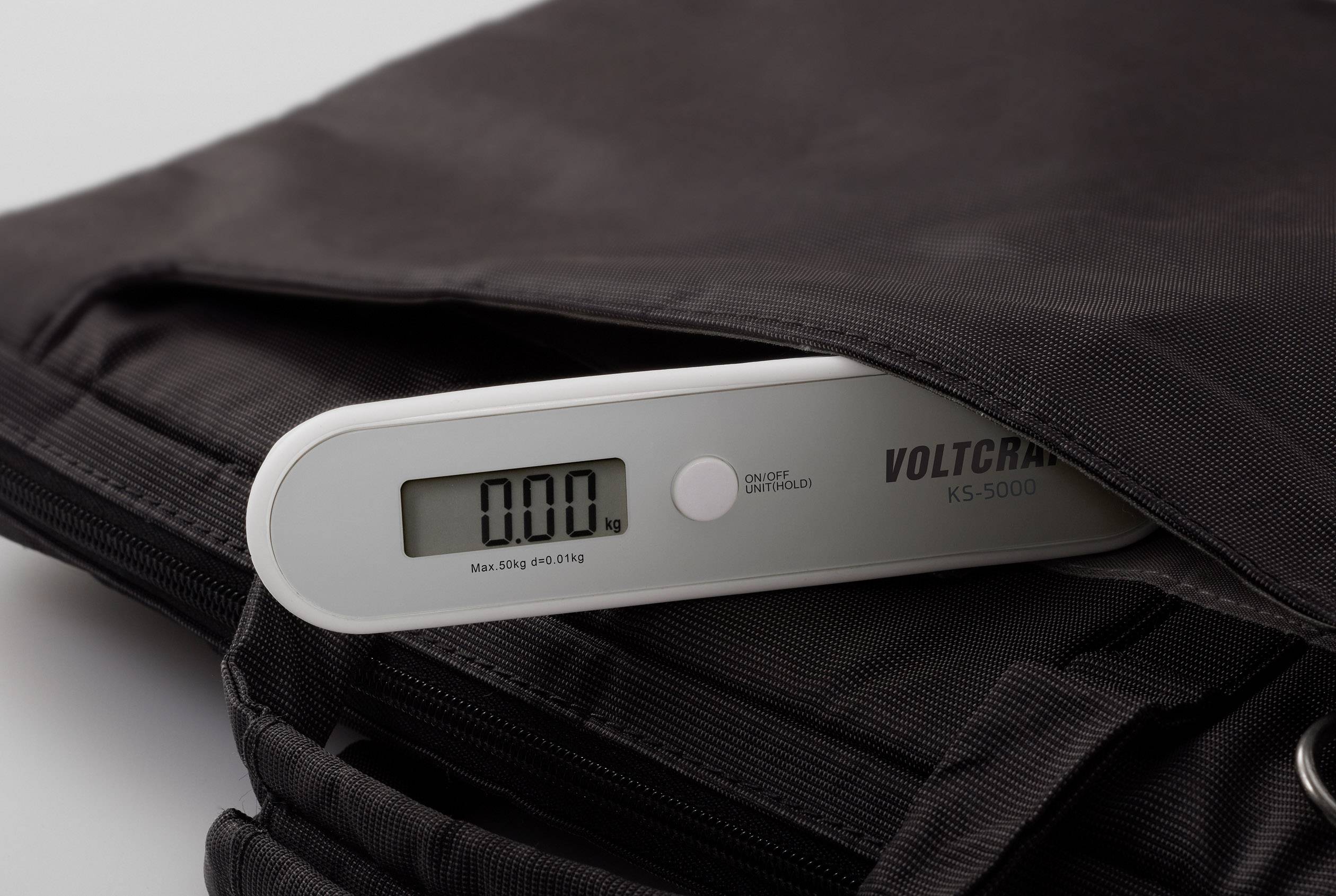 'Digitale Gepäckwaage mit Display zeigt 0,00 kg an. Gerät liegt auf einer schwarzen Tasche. Markenname 'Voltcraft' sichtbar.'