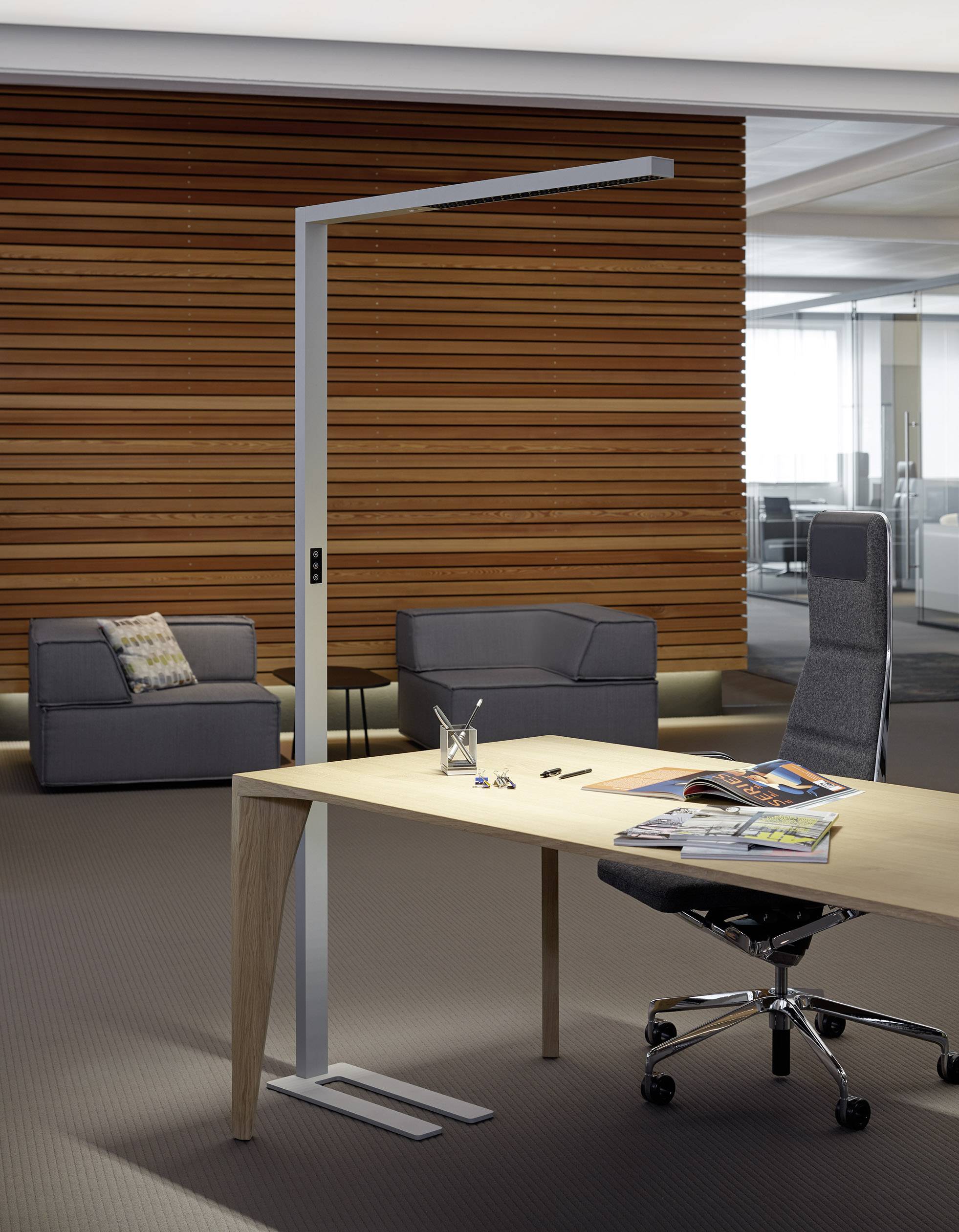 Moderner Büroraum mit Holzwand: Schreibtisch mit hochwertigen Stuhl und Stehleuchte, zwei graue Sessel im Hintergrund, Akten auf dem Tisch.