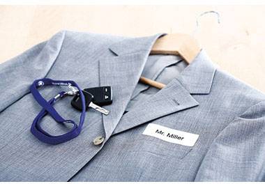 Grauer Anzug mit Namensschild 'Mr. Miller', Schlüsselbund und blauer Schlüsselband auf Holzkleiderbügel.