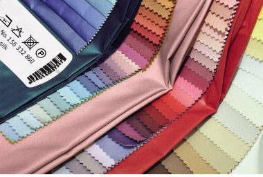 Stoffmuster in verschiedenen Farben, von Blau über Rosa bis Rot, Fächerartig angeordnet, mit Etikett auf der linken Seite.