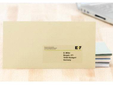 Ein gelber Briefumschlag mit der Aufschrift 'D. Müller, Bergerstr. 27/1, 70184 Stuttgart, Germany', liegt auf einem Tisch. Im Hintergrund ein Laptop.