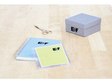 Ein spiralgebundenes Notizbuch, eine CD-Hülle und eine Schachtel mit 'Elf'-Logo auf einem Holztisch, daneben eine Schere.