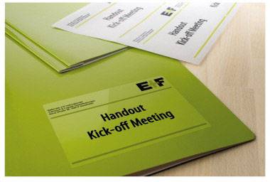 Grüne Mappe mit Aufschrift 'Handout Kick-off Meeting'. Weitere Blätter und Firmennamen darauf sichtbar. Bürosetting.