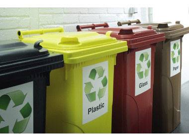 Mehrere farbige Mülltonnen mit Recycling-Symbolen und Beschriftungen: 'Plastic', 'Glass', 'Paper', aufgereiht vor einer Wand.