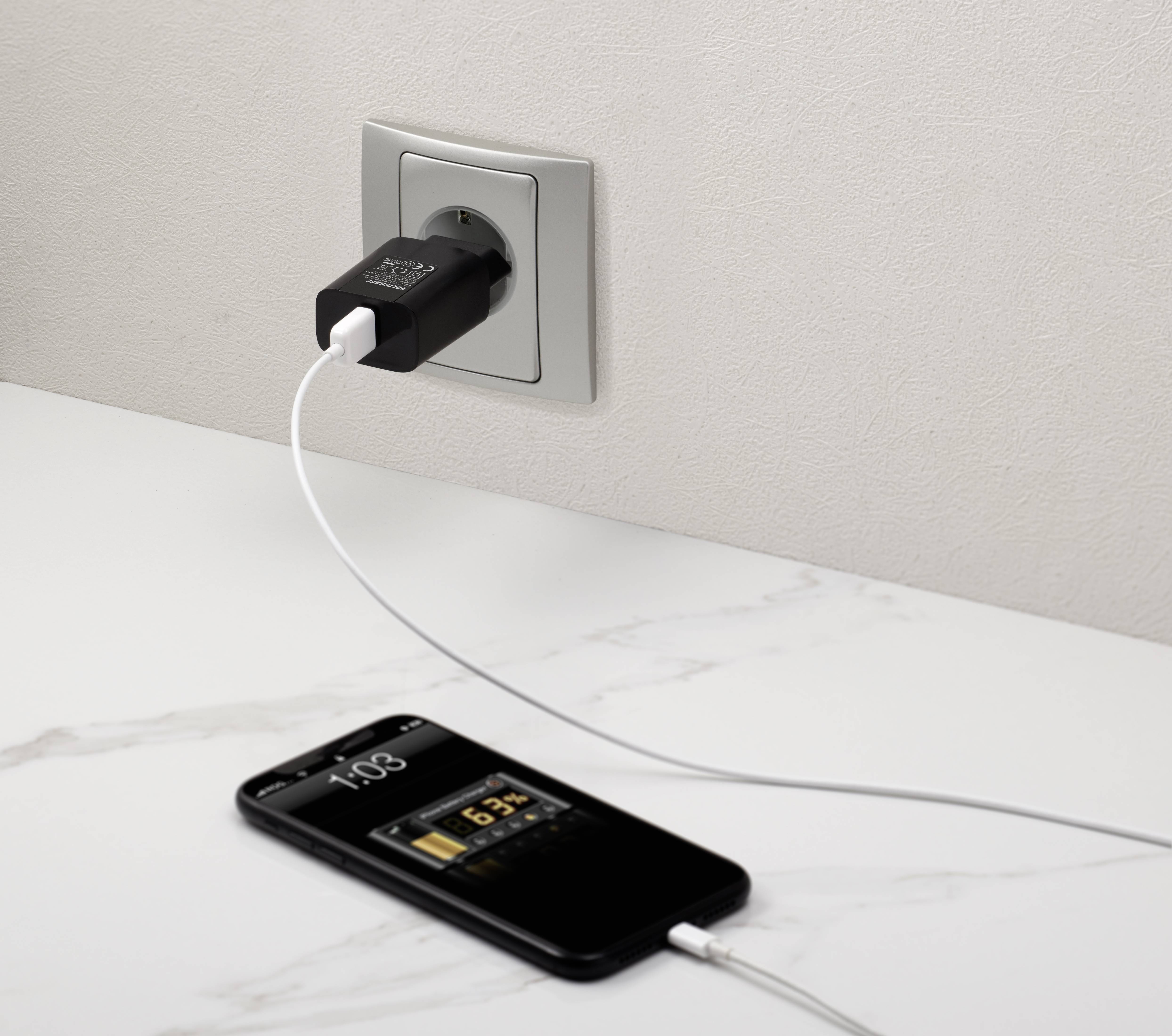 Ein Smartphone wird über ein USB-Kabel an einer Steckdose aufgeladen. Das Display zeigt '65%' und die Uhrzeit an.