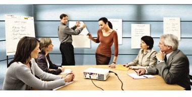 Sechs Personen in einem Meetingraum diskutieren vor einer Tafel. Zwei Personen stehen und schreiben, während vier am Tisch sitzen. Auf dem Tisch steht ein Projektor.