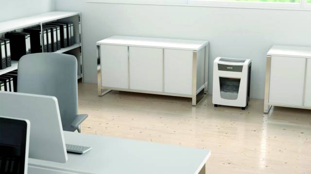 Ein helles Büro mit Holzfußboden, einem Schreibtisch, einem Regal mit Ordnern und einem Aktenvernichter in der Ecke.