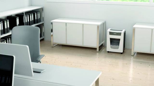 Ein moderner Büroraum mit einem Aktenschredder auf dem Boden. Schreibtisch mit Computer im Vordergrund, Ordner in Regalen.