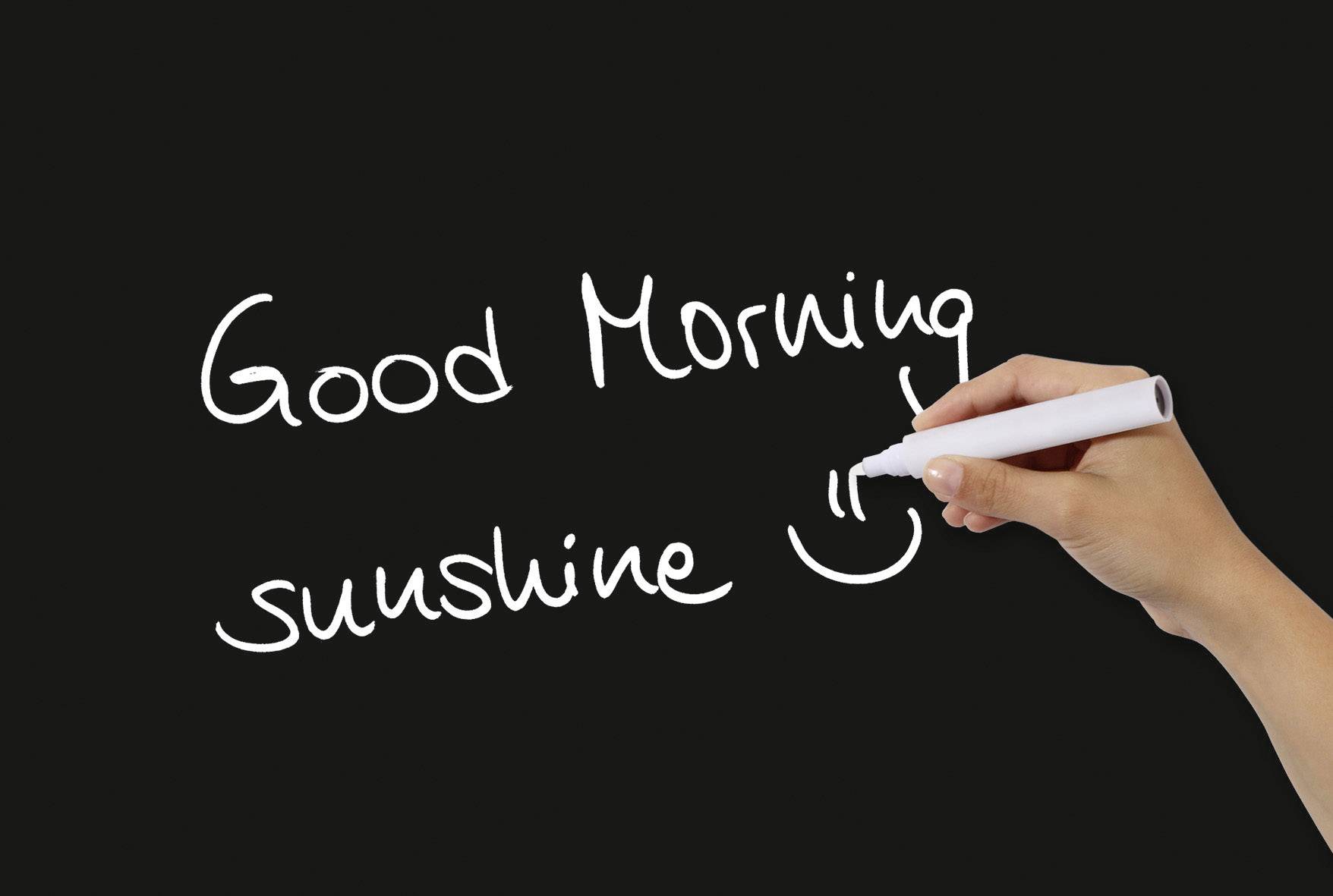 Eine Hand schreibt mit einem weißen Stift auf eine schwarze Tafel: 'Good Morning sunshine', ergänzt durch ein lächelndes Gesicht.
