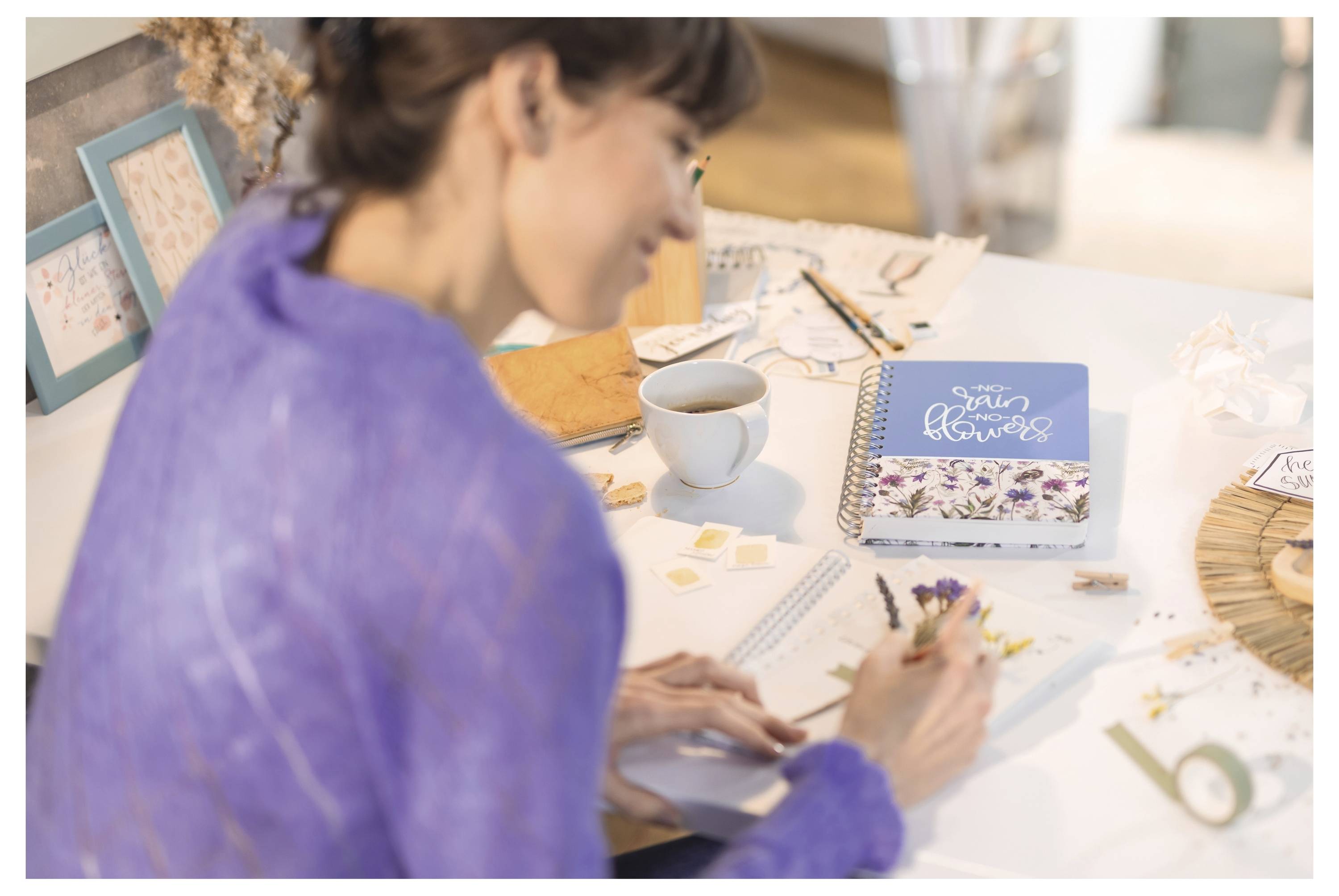Frau in einem violetten Oberteil, die an einem Basteltisch in ein Notizbuch schreibt, mit einer Kaffeetasse, einem Blumennotizbuch, Stiften und Bastelmaterialien.