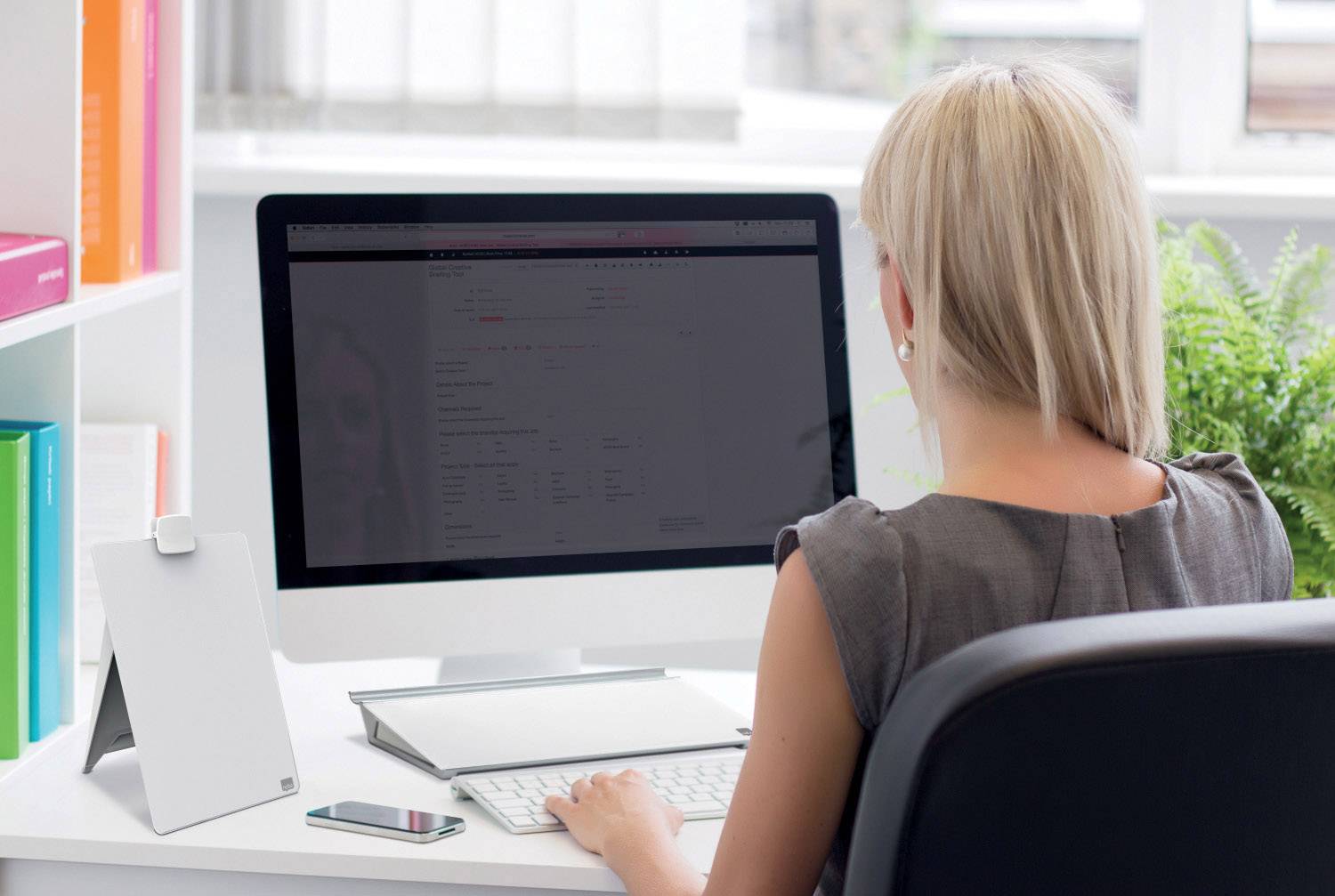 Eine Frau sitzt an einem Schreibtisch und arbeitet an einem Computer. Ein Smartphone liegt daneben. Im Hintergrund sind Pflanzen zu sehen.
