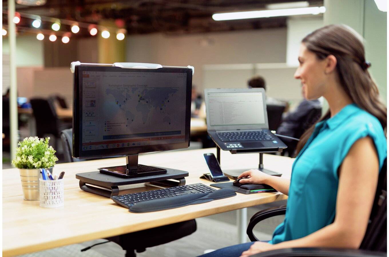 Eine Frau sitzt in einem modernen Büro an einem Schreibtisch mit zwei Monitoren, einem Laptop und einem Smartphone. Sie arbeitet konzentriert am Computer.