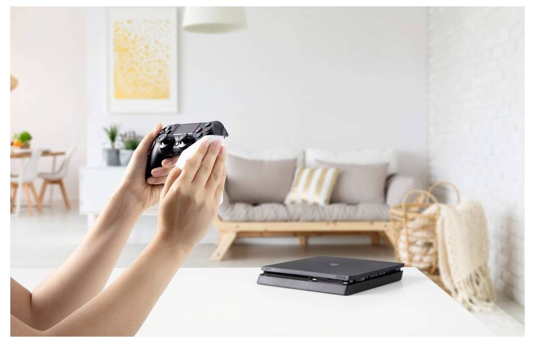 Eine Person desinfiziert einen Videospielcontroller mit einem Tuch in einem hellen Wohnzimmer, mit einer Konsole auf dem Tisch und einem Sofa im Hintergrund.