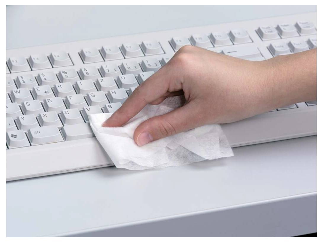 Eine Hand reinigt eine Computertastatur mit einem weißen Desinfektionstuch und betont damit Hygiene und Wartung in einer Büroumgebung.