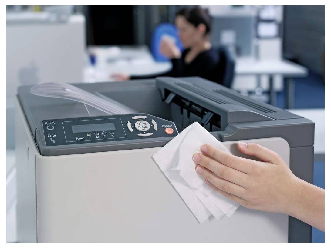 Eine Person reinigt einen Bürodrucker mit einem Tuch; ein unscharfer Büroangestellter ist im Hintergrund zu sehen.
