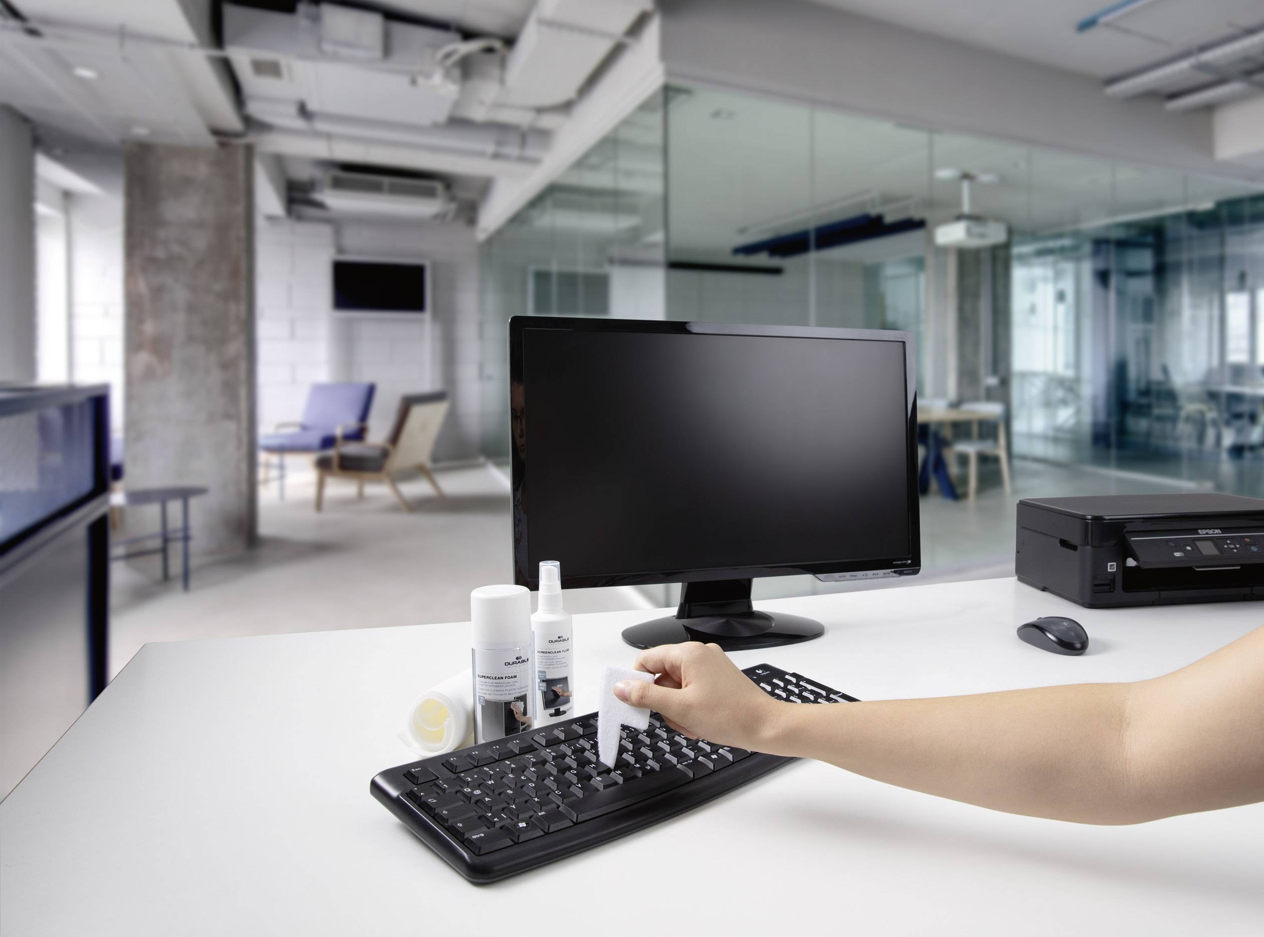 Eine Person reinigt eine Computertastatur in einem modernen Büro mit Reinigungsspray und Tuch. Im Hintergrund sind Glaswände und Bürostühle zu sehen.