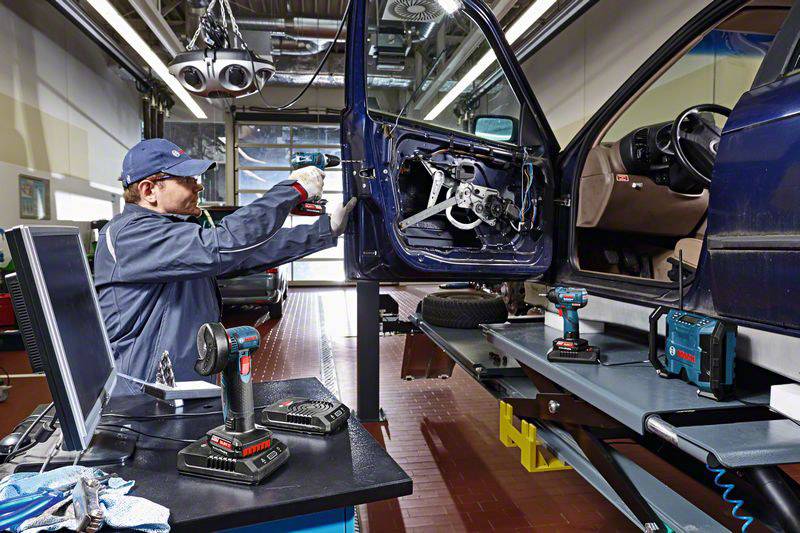 Ein Mechaniker arbeitet in einer Werkstatt an der Fahrertür eines aufgebockten Autos. Verschiedene Werkzeuge sind auf einem Tisch.