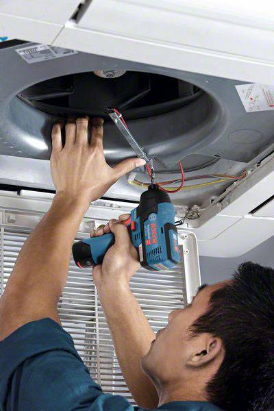 Eine Person repariert eine Klimaanlage an der Decke mit einem blauen Akkuschrauber; Kabel sind sichtbar.