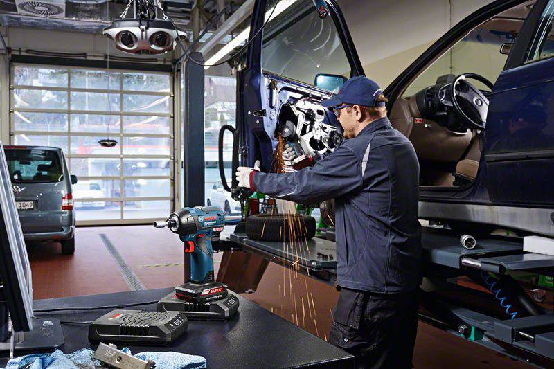 Ein Mechaniker bearbeitet mit einem Werkzeug die Tür eines Fahrzeugs in einer Werkstatt. Funken sprühen vom Metall. Im Hintergrund stehen weitere Autos.