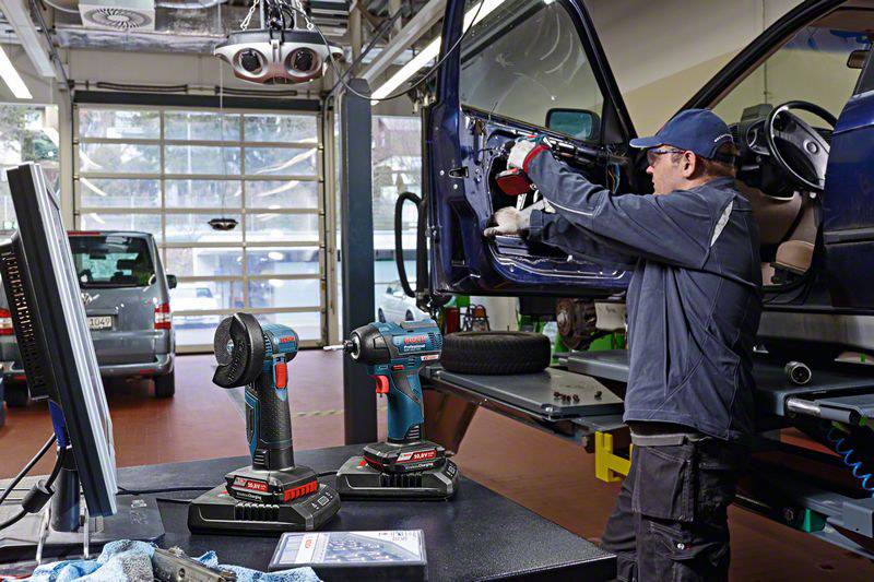 Ein Mechaniker in einer Werkstatt repariert das Armaturenbrett eines Autos. Zwei Akkuschrauber stehen im Vordergrund auf einem Tisch.