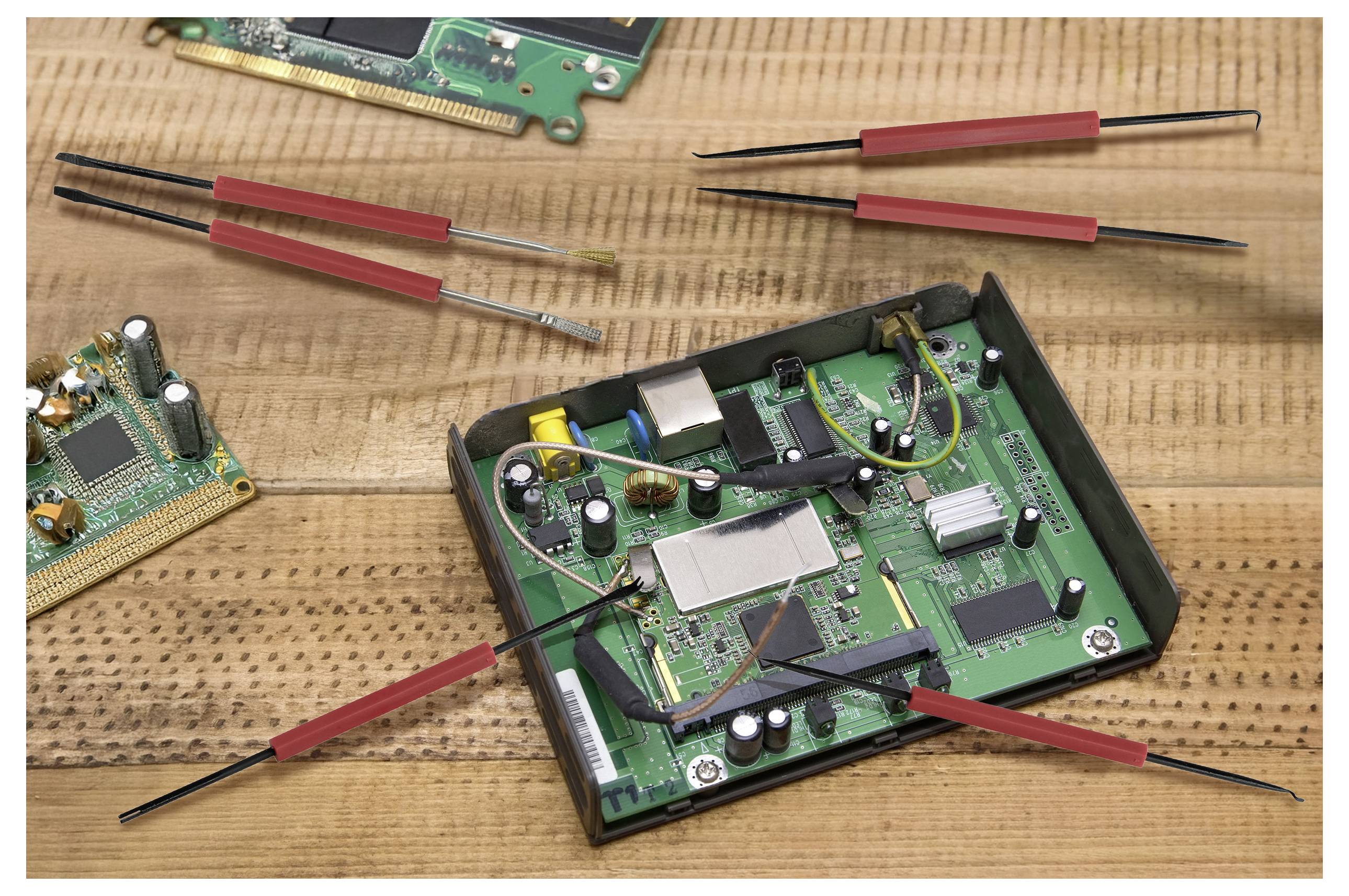 Eine offene Leiterplatte auf einem Tisch mit elektronischen Komponenten. Mehrere rote und schwarze Pinzetten sind drum herum verteilt, was auf Reparaturarbeiten hindeutet.
