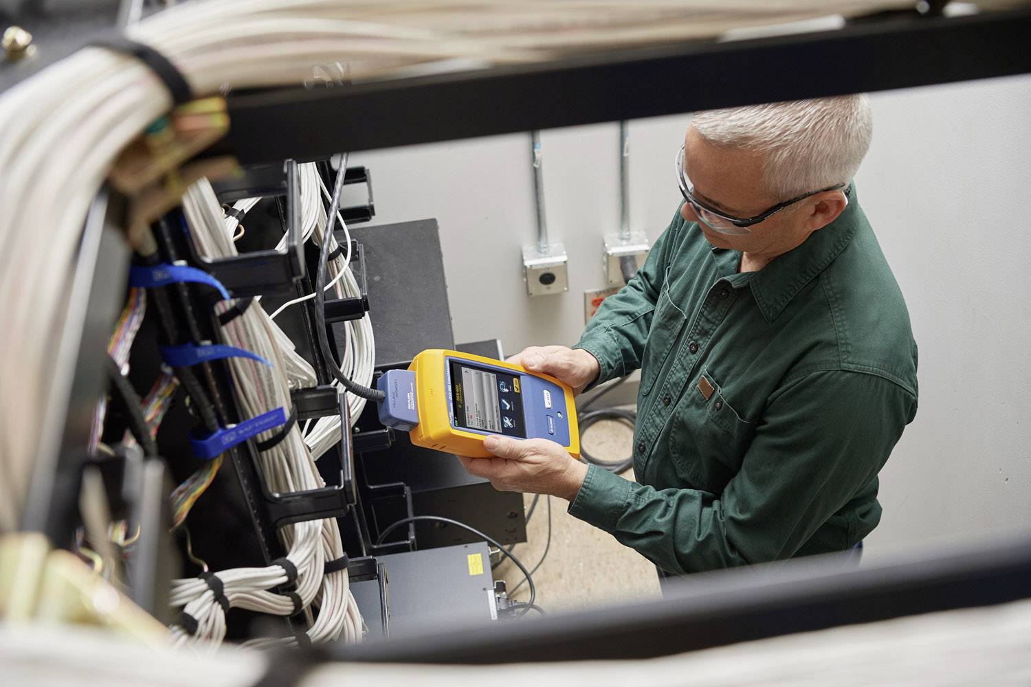 Eine Person prüft mit einem gelben Gerät Netzwerkkabel in einem Serverraum. Sie trägt eine grüne Jacke und eine Brille.