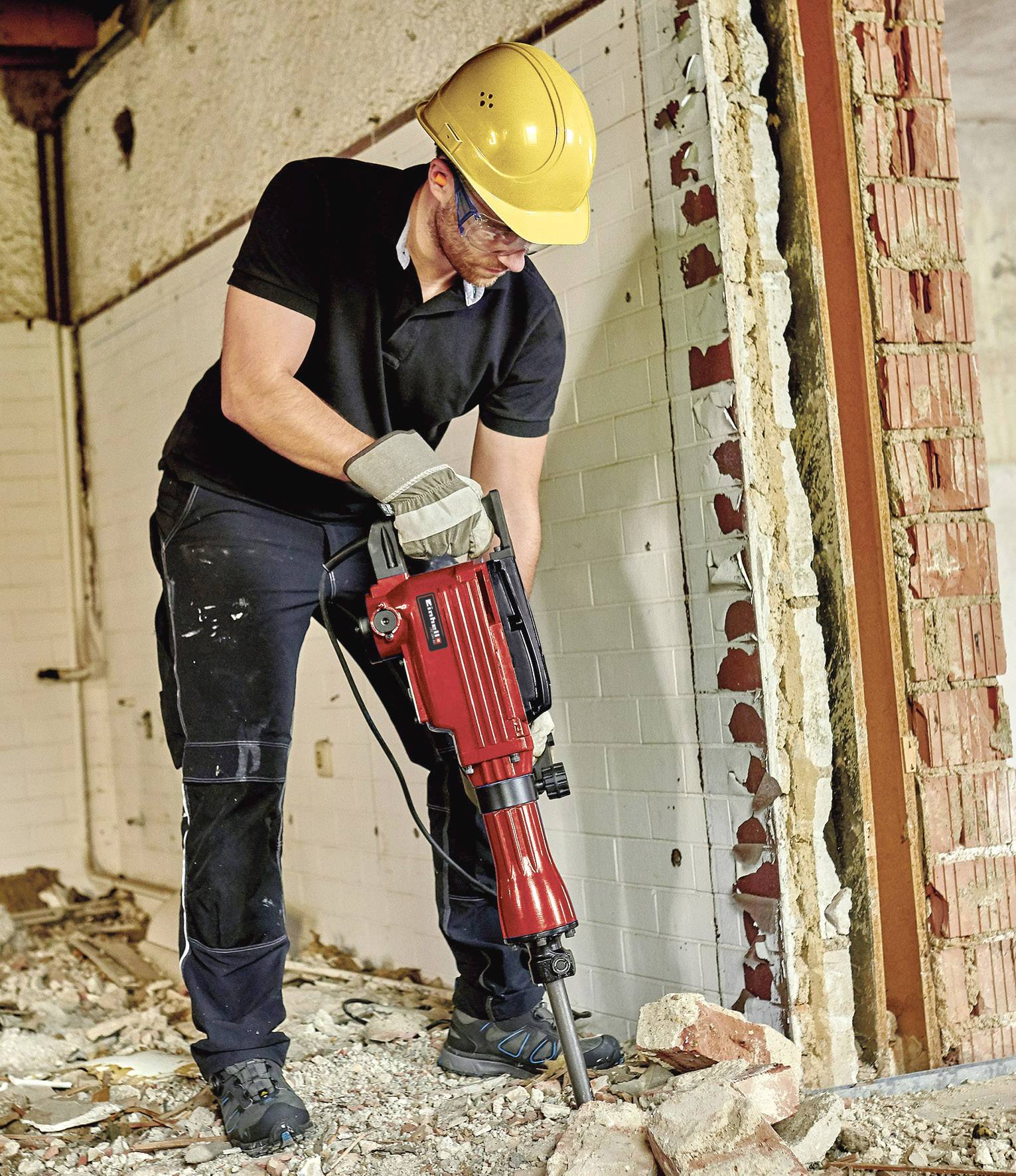 Ein Bauarbeiter mit Schutzhelm und Handschuhen verwendet einen roten Presslufthammer, um eine Backsteinwand in einem Gebäude zu bearbeiten.